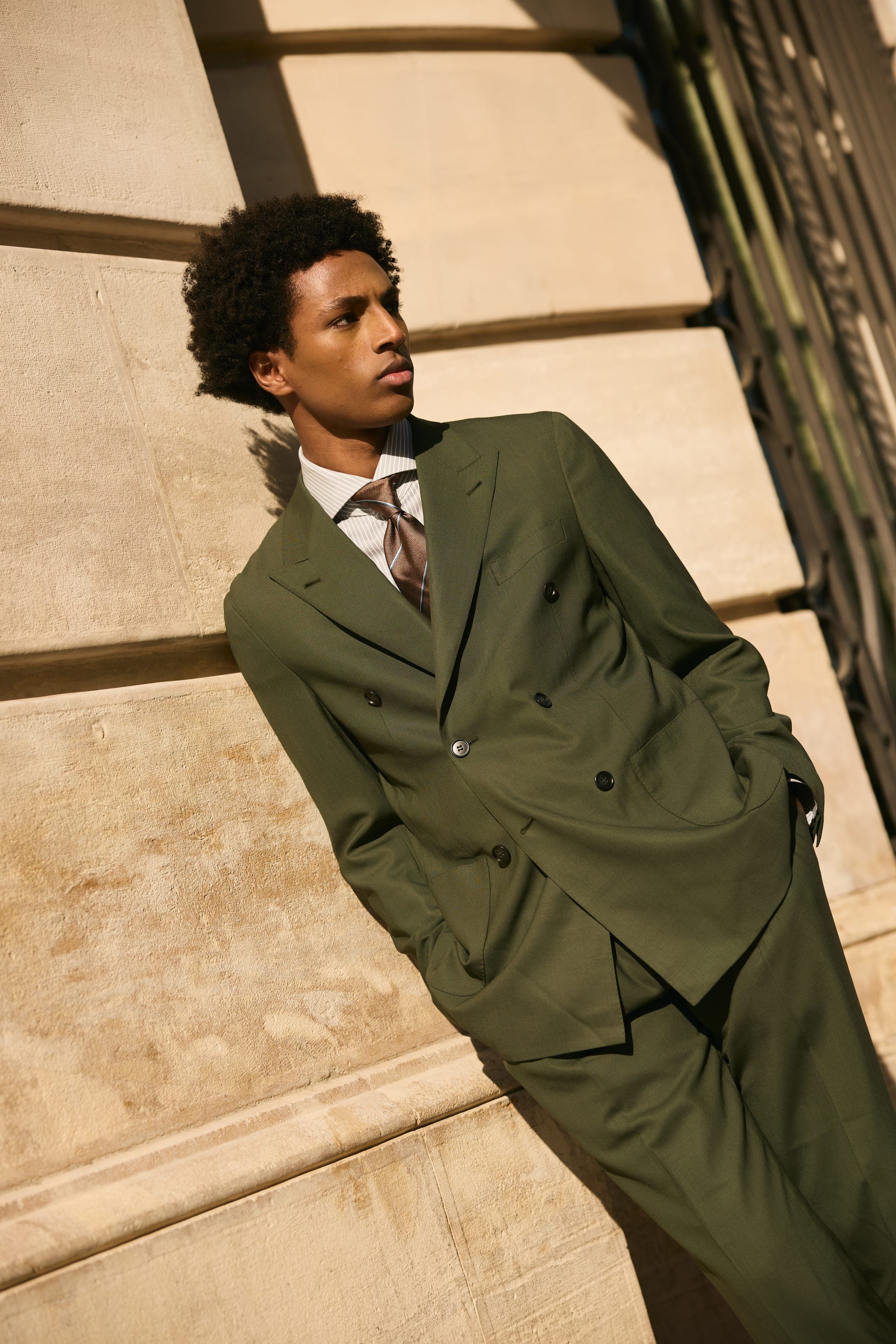 A tailored olive suit against a sunlit stone wall